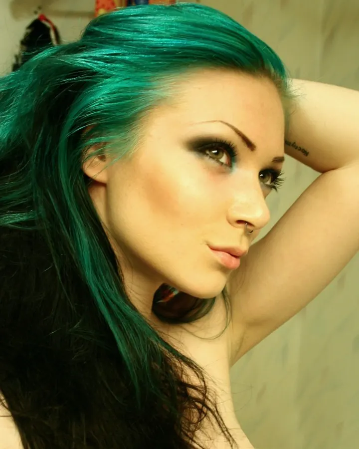 Femme aux cheveux verts et maquillage sombre, regardant de côté, bras levé derrière la tête