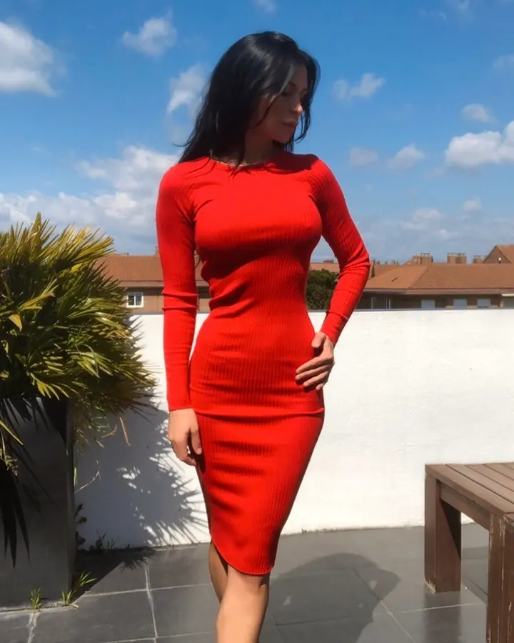 Femme en robe noire, debout près d’un mur, souriant légèrement