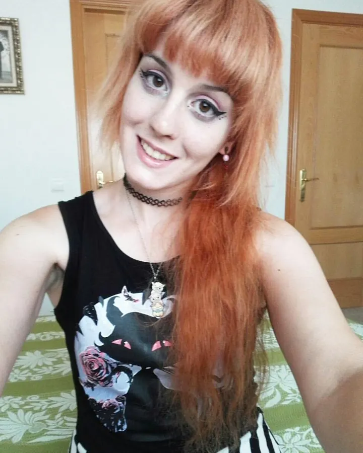 Femme aux cheveux roux, en haut noir sans manches avec un motif de chat, souriant à l'appareil photo dans une pièce intérieure.