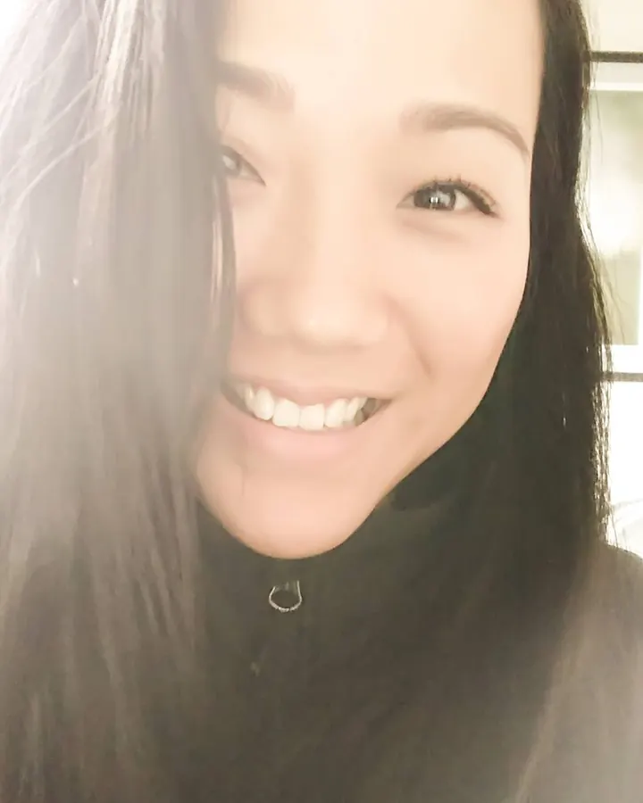 Femme asiatique aux cheveux noirs, vêtue d'un haut noir, assise sur un lit, dos tourné, regard vers l'appareil, cheveux mouillés.