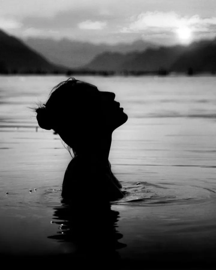 Silhouette d'une femme dans l'eau, face au soleil couchant, en noir et blanc.