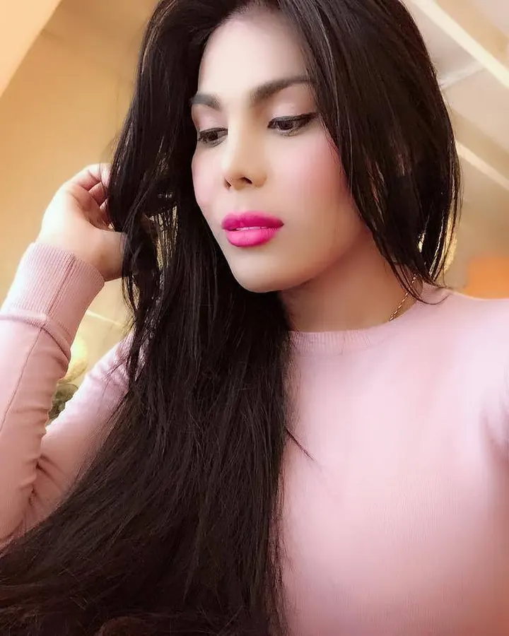 Femme aux cheveux noirs longs portant un pull rose, regard baissé, main dans les cheveux.
