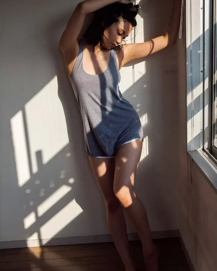 Femme aux cheveux noirs portant un body gris, posant contre un mur blanc avec des rayons de soleil traversant la fenêtre.