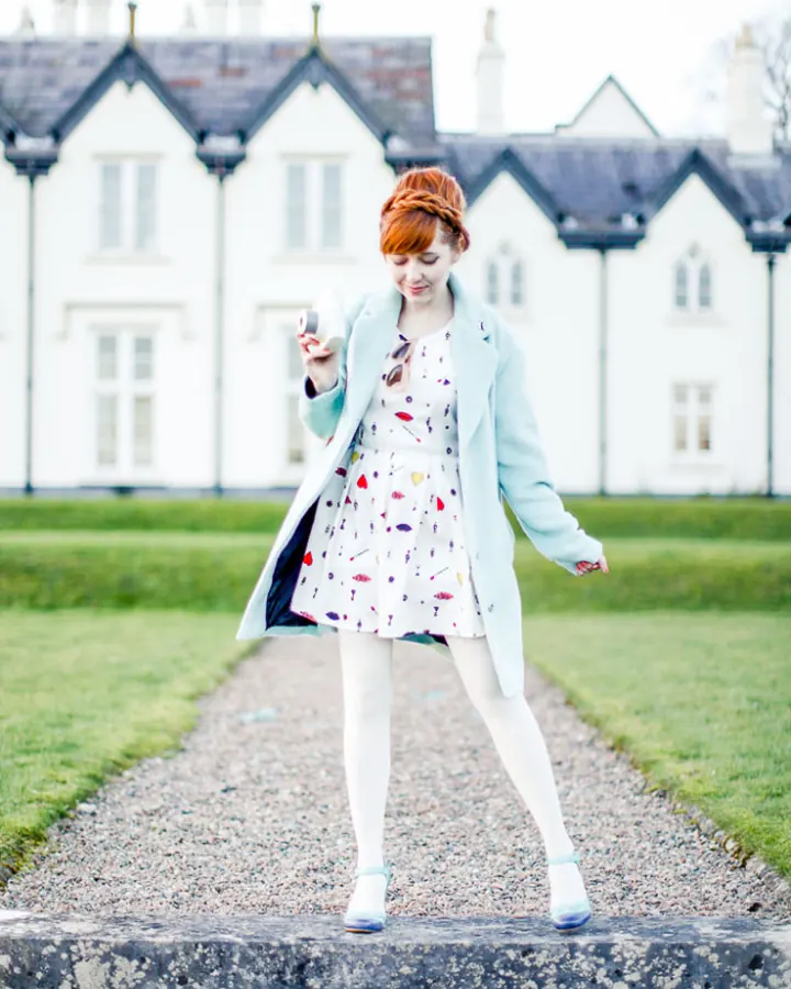 Femme aux cheveux roux, vêtue d'une robe légère et d'un manteau bleu ciel, marchant sur un chemin pavé devant une grande maison blanche.
