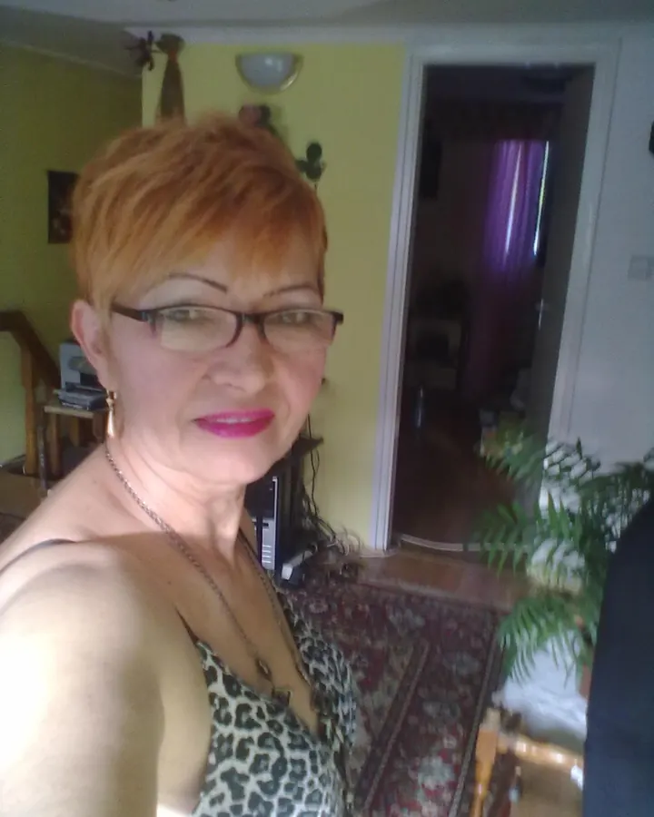 Femme aux cheveux bruns foncés, portant un haut noir, une jupe en cuir synthétique noire, des lunettes, debout dans un couloir, regardant vers le bas.