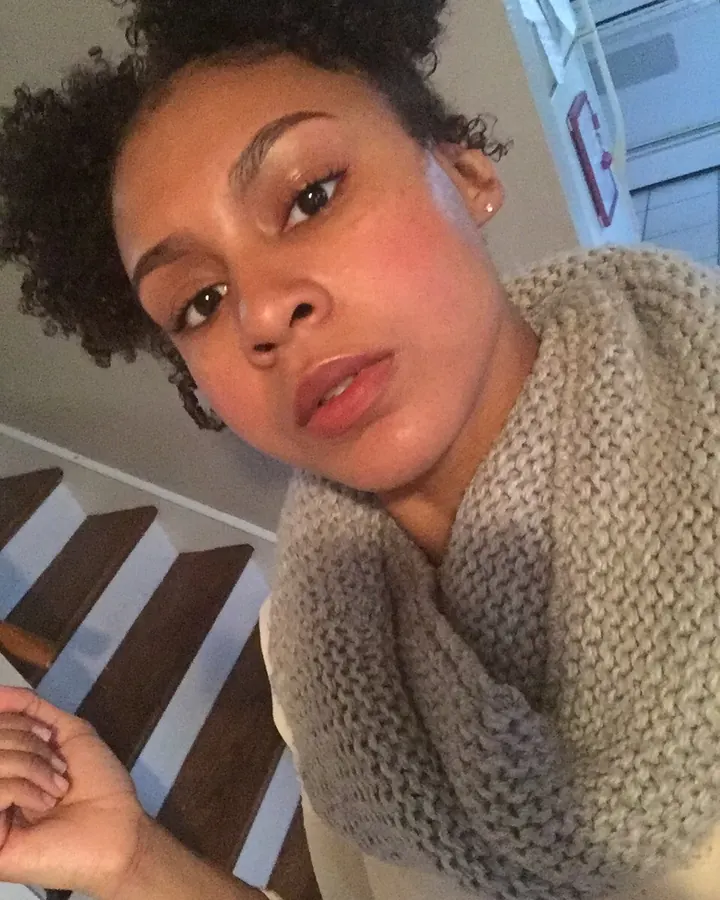 Femme aux cheveux bruns noirs portant un haut noir, posant dans une chambre avec un lit en arrière-plan