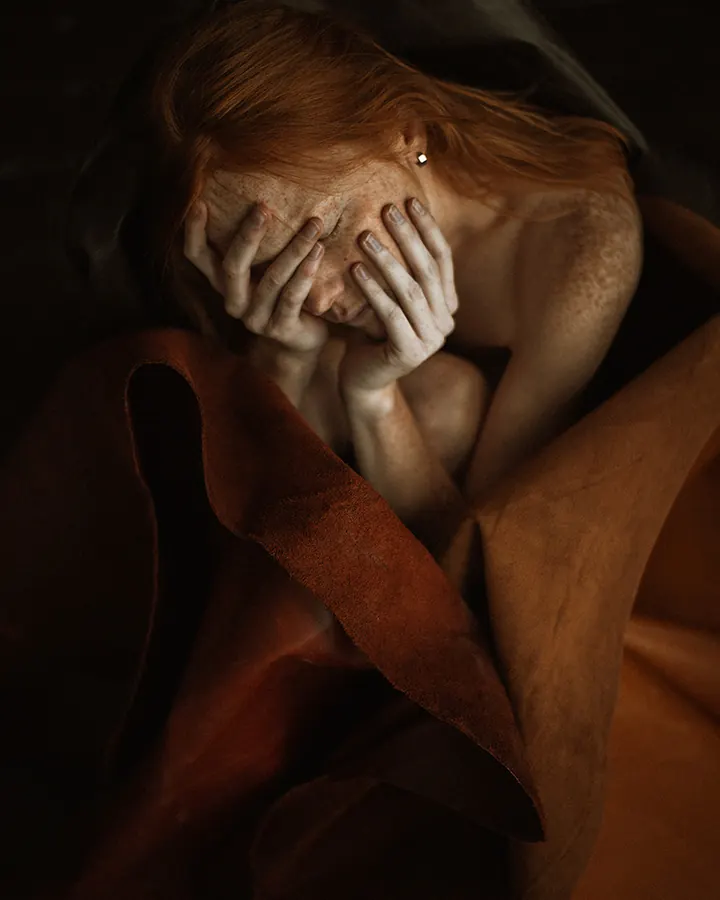 Femme aux cheveux roux cachant son visage avec ses mains, enveloppée dans une couverture marron, dans une ambiance sombre.